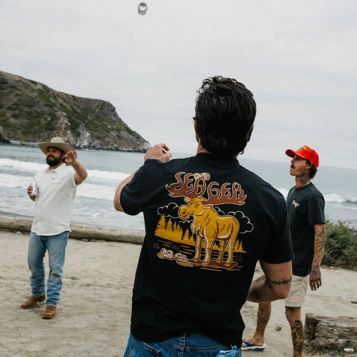 Men wearing the Tipsy Moose graphic tee on the beach, tossing a drink, casual rugged style shirt for men